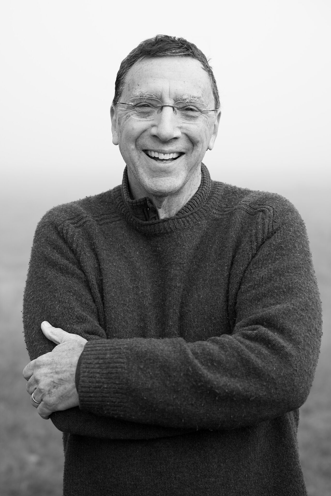 John Markoff in 2022, in a black and white photo taken by Christopher Michel, crossing his arms in a thick woolly jumper and smiling, in front of a neutral background.